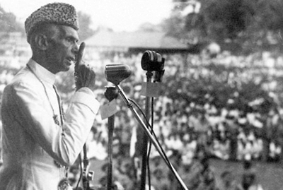 A speaker in white attire addresses a large crowd at an outdoor gathering, with microphones set up for amplification.