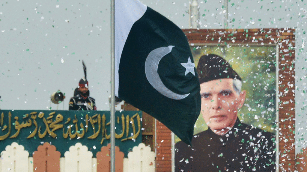 A Pakistani flag waves amidst green and white confetti, with a soldier standing guard behind a decorative banner and a large portrait.